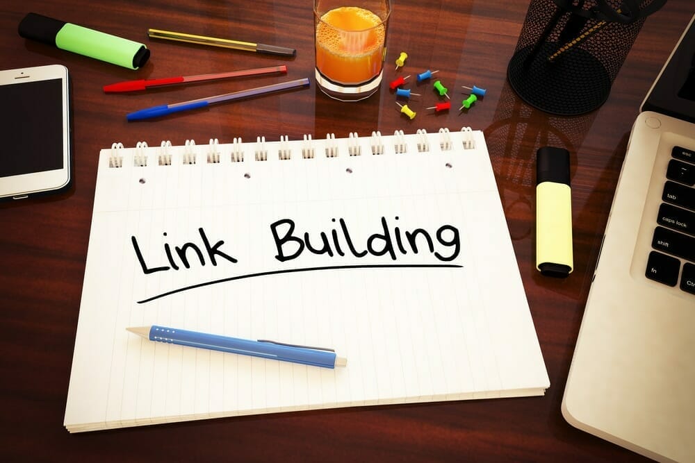 The words "Link Building" written on a notebook that is sitting on a desk with a computer and smartphone.