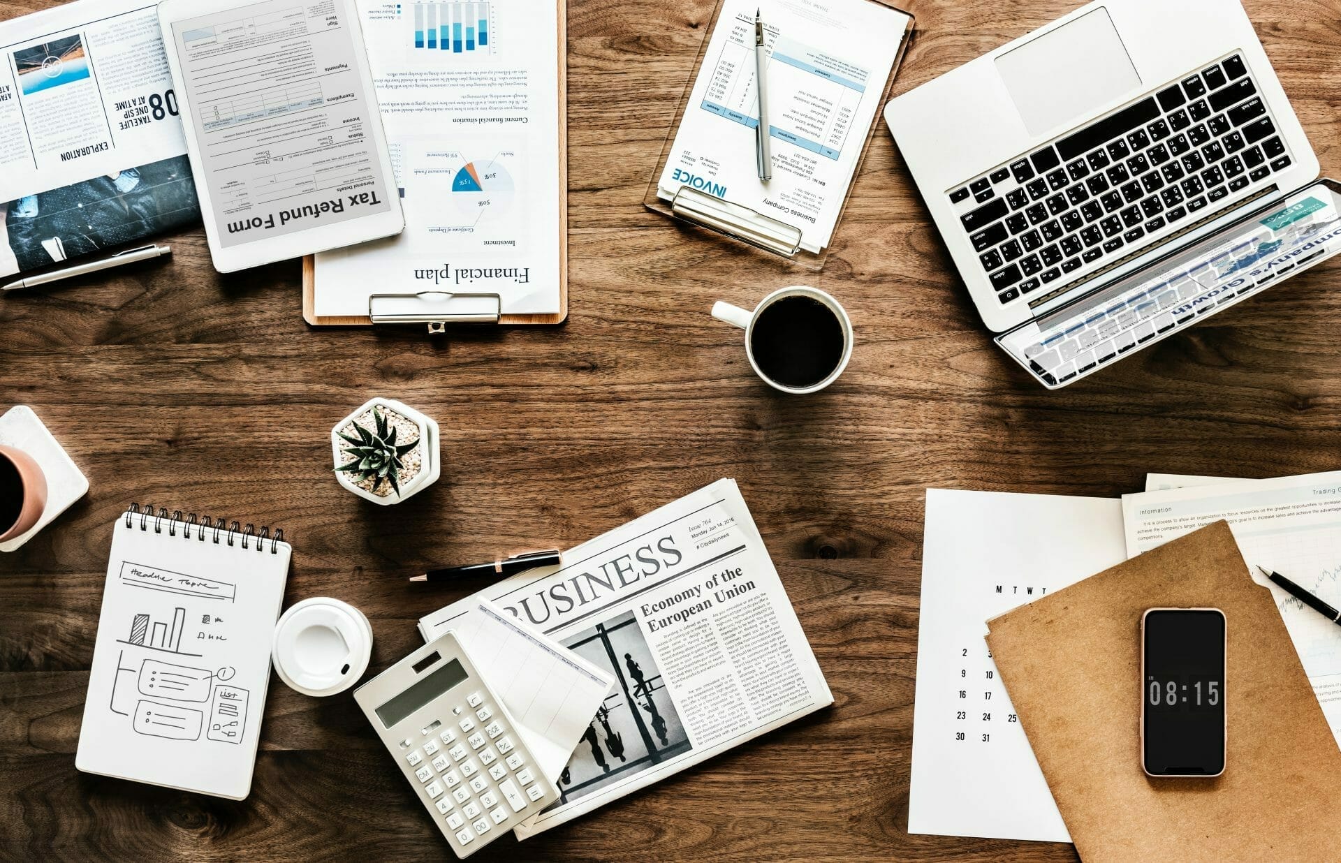 Tools for conducting business on a wooden desk including a laptop, smartphone, clipboard, newspaper, calendar, and coffee.