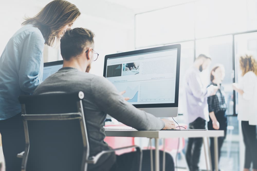 Woman looking over the shoulder of a man seated at a large computer screen