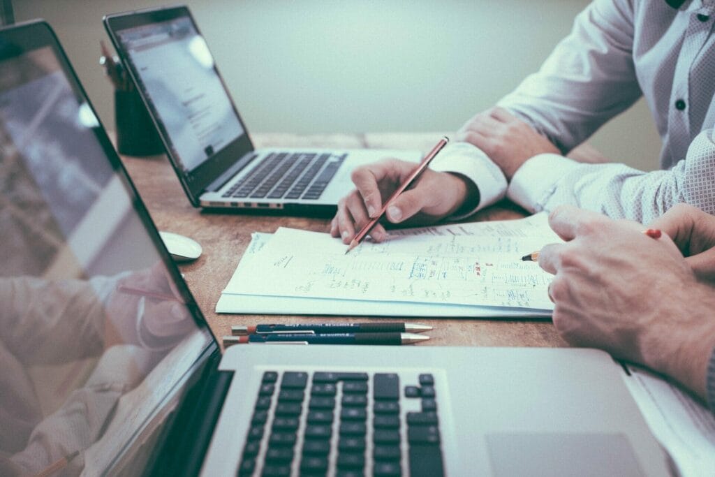 Two men at a table going over paperwork with 2 laptops nearby