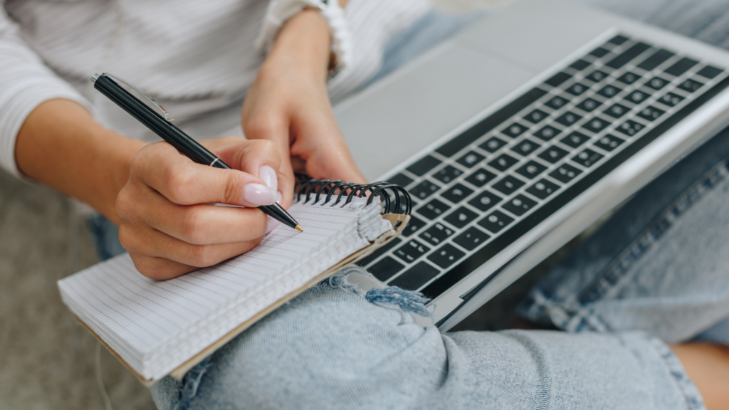 Person writing in a notepad with laptop computer on their lap