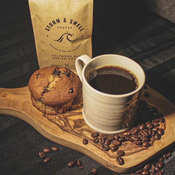 Photo of a bag of Storm & Swell coffee on a wooden serving platter with a muffin, a cup of coffee, and roasted coffee beans.