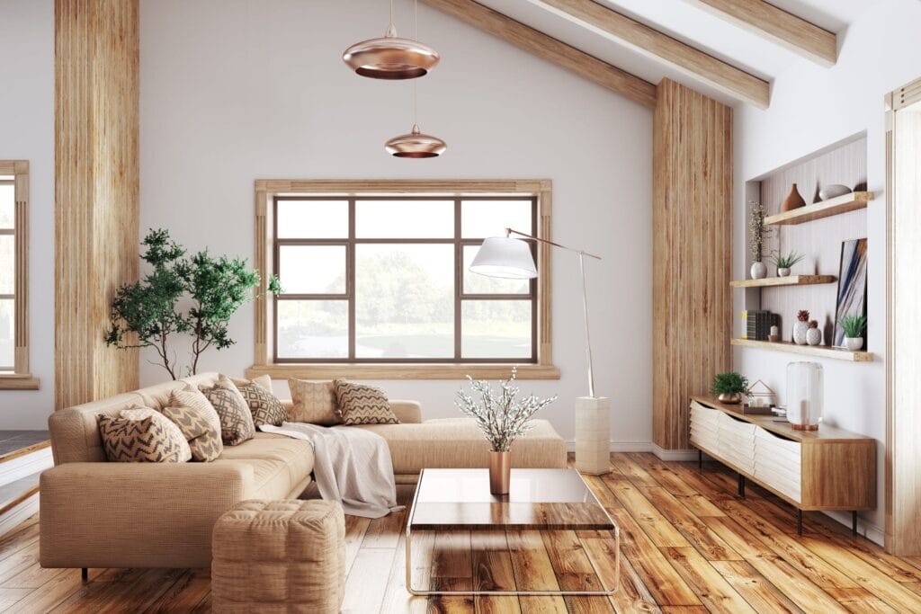 Modern living room with a beige L-shaped sofa, wooden flooring, and large windows. Features include a wooden coffee table, indoor plants, wall-mounted shelves with decor, and slanted ceiling with hanging pendant lights. Natural light fills the space.