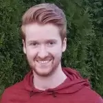 A person with red hair, a beard, and wearing a red hoodie smiles while standing in front of a leafy green background.