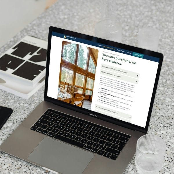 A laptop on a speckled countertop displays a website featuring an interior cabin image and text about vacation rentals. Next to the laptop are clear plastic cups and a color swatch book.
