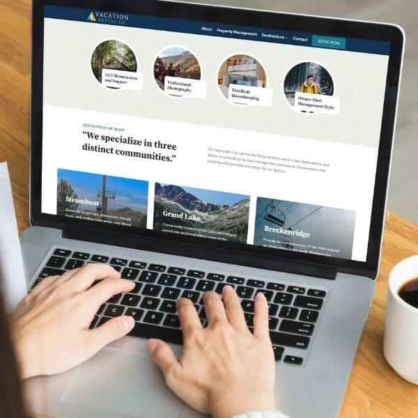 Person typing on a laptop displaying a vacation rental website. The screen shows navigation options and features like 24/7 maintenance, professional cleaning, and guest-first management. A coffee cup is beside the laptop.