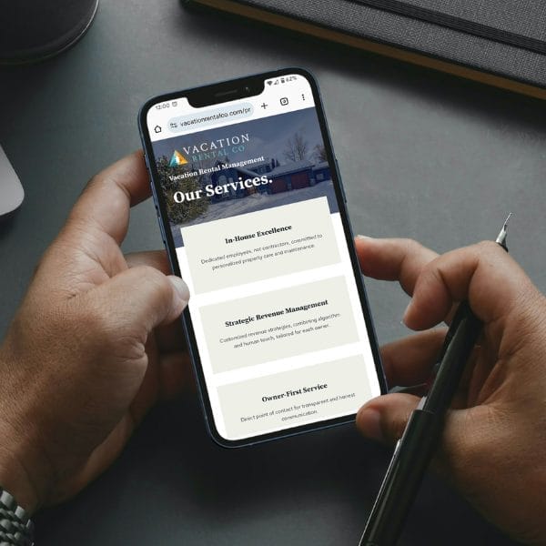 A person holds a smartphone displaying a website about vacation rental management. The screen shows services such as in-home excellence, strategic revenue management, and owner-first service. A pen is held in the other hand.