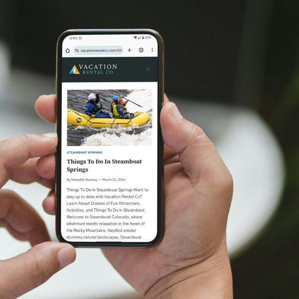 A person holds a smartphone displaying an article about things to do in Steamboat Springs on the Vacation Rental Co. website. The screen shows a header image of a group rafting. The phone is held with one hand over a blurred background.