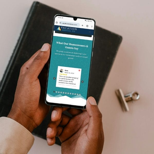 A person holds a smartphone displaying a website for "Vacation Rental Co" with a section titled "What Our Homeowners & Guests Say." The phone is held over a dark book on a table. A metal clip is nearby.