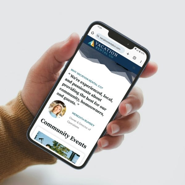 Hand holding a smartphone displaying a website for "Vacation Rental Co." The screen shows text about community events and a small image of a woman labeled as "Community Director." The website's header is visible at the top.