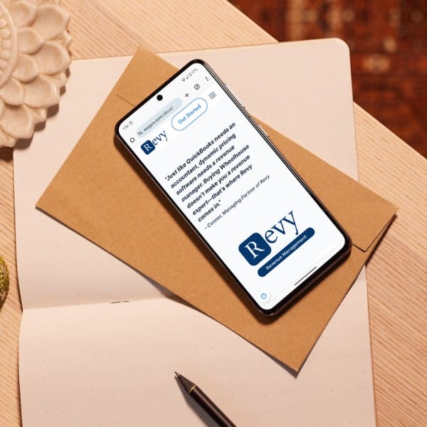 Smartphone displaying a webpage on top of an open notebook and a brown envelope. The notebook has dotted pages. In the background, a white decorative object and part of a rug are visible.