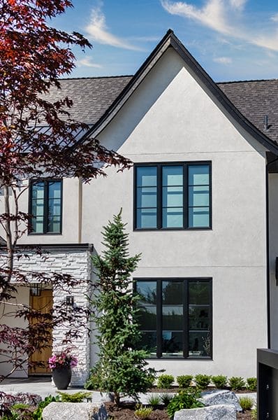 A modern two-story house with a pitched roof and large black-framed windows is surrounded by landscaped greenery. A tall pine tree and a red-leaved tree are in the foreground under a blue sky with wispy clouds.