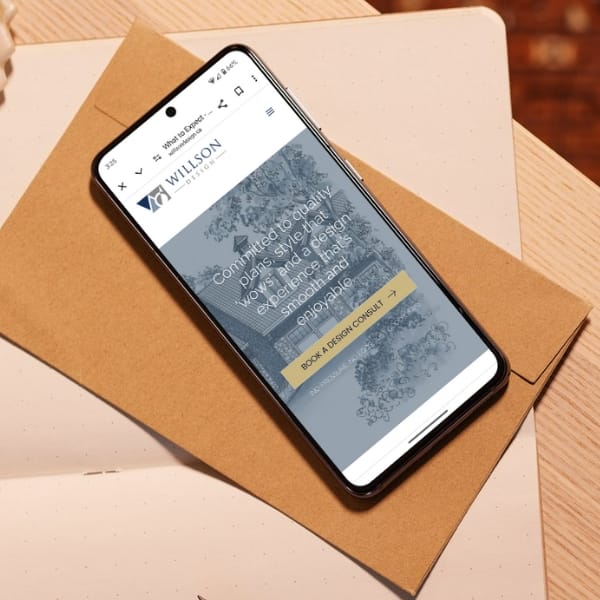 A smartphone displaying the Willson website rests on two brown envelopes on a table. The screen shows text promoting originality and high-standard workspace design. The phone is placed at an angle, and the background is softly blurred.