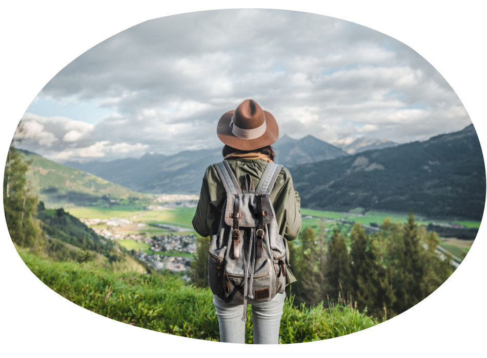 A person wearing a hat and backpack stands on a grassy hill, overlooking a scenic valley with mountains, greenery, and a cloudy sky in the background.
