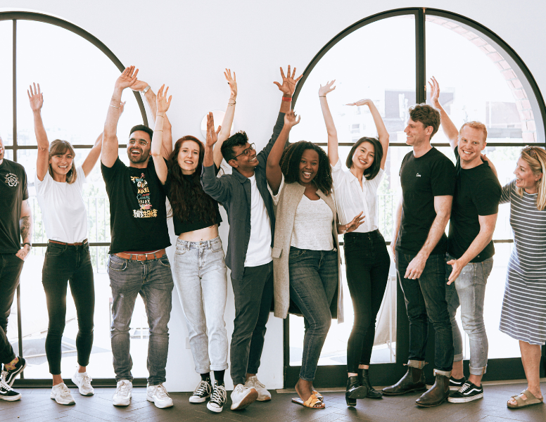 A diverse group of people stands in a line, smiling and raising their hands in a celebratory pose. They are standing indoors in front of large arched windows. Everyone is casually dressed, creating a joyful and relaxed atmosphere.