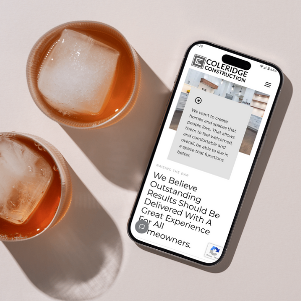 A smartphone displaying the Cole Ridge Construction website is placed on a light-colored surface next to two iced coffee glasses. The website shows text about creating homes with outstanding results.