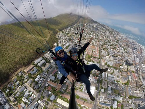 Two people tandem paragliding high above a cityscape. Both wear helmets and harnesses, with one holding a selfie stick. Green hills are visible in the background, and the city stretches out below under a blue sky with scattered clouds.