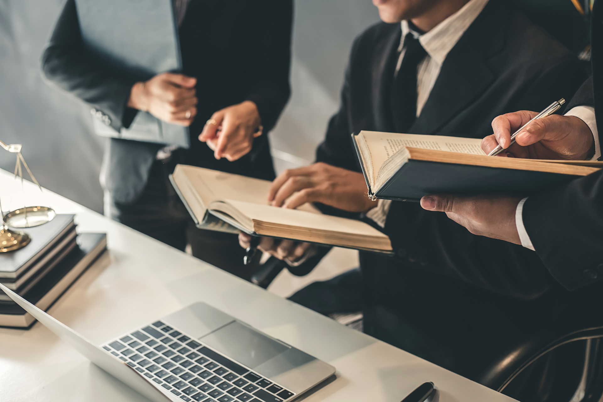 Three people in business attire are gathered around a table with a laptop, holding and discussing open books. A set of scales and more books are on the table, suggesting a legal or professional meeting.
