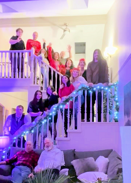 A group of people gathered on a staircase and a couch, smiling and waving at the camera. The staircase is decorated with blue and white lights, and a person stands at the top. The setting is indoors with warm lighting.