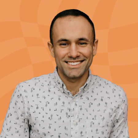 A person with short dark hair, smiling and wearing a light-colored button-up shirt with small patterns, stands in front of an orange wavy background.