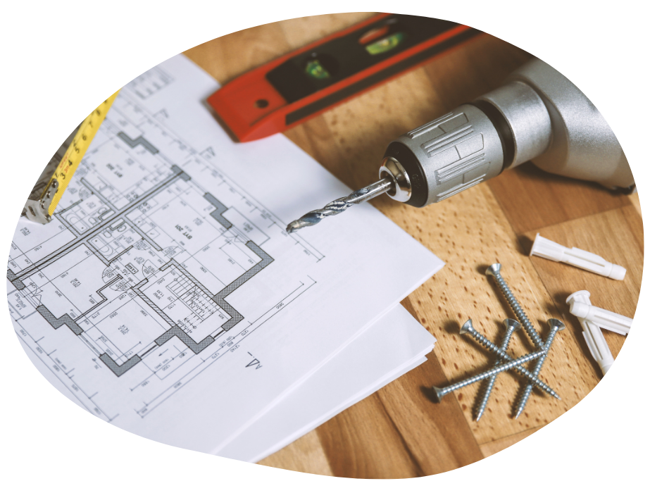 A drill, measuring tape, and spirit level are placed on a wooden surface alongside screws and wall plugs. Blueprints of a floor plan lay beneath them, suggesting a construction or renovation setting.