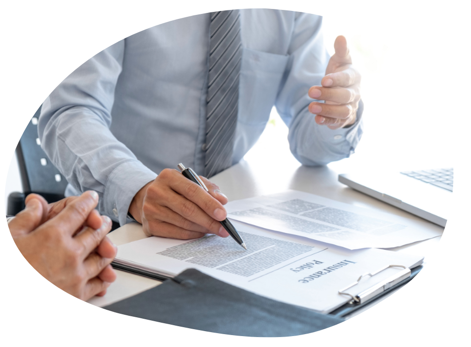 Two people in business attire sit at a desk, reviewing documents labeled "Insurance Policy." One person gestures while the other points at the papers with a pen; a laptop and calculator are also visible.