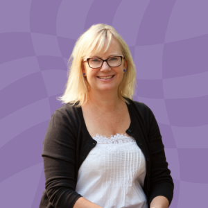A smiling woman with blonde hair and glasses, wearing a white top and black cardigan, sits in front of a purple wavy-patterned background.